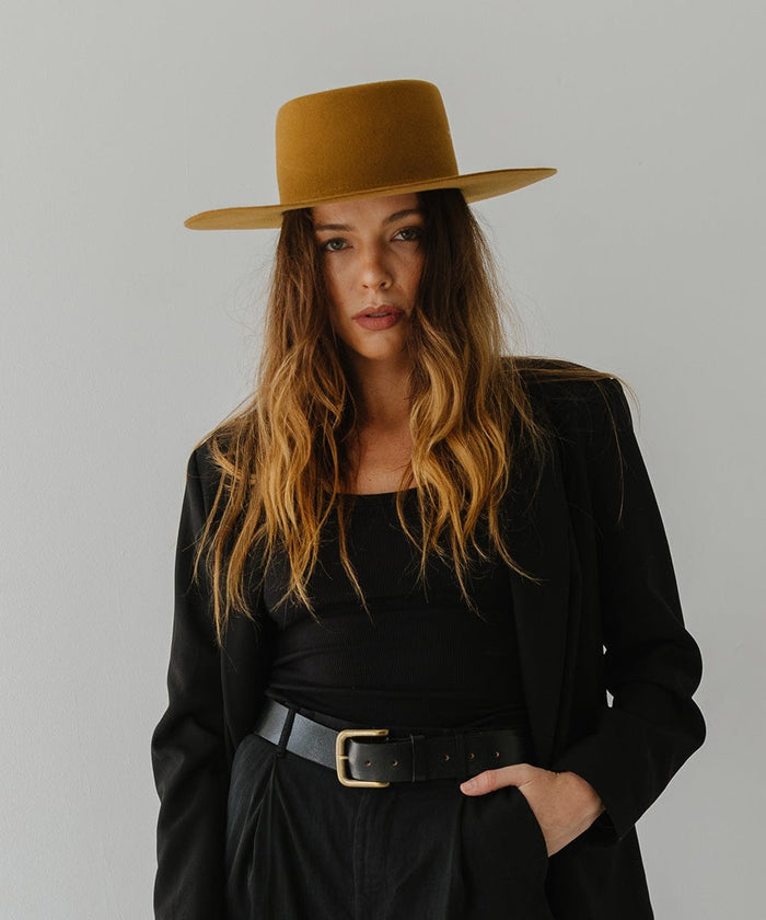 Woman wearing a cinnamon colored boater hat, a black top with a black coat, black belt and black pants against a plain background #color_cinnamon