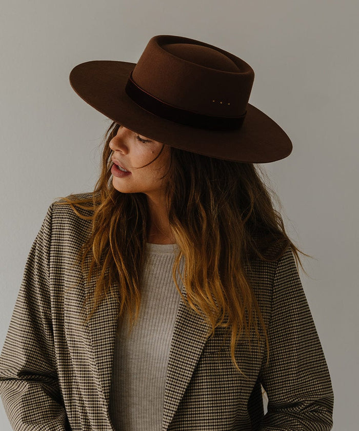 Woman wearing a dark oak colored boater hat with a hat band and white top with a coat, looking sideways against a plain background #color_dark oak