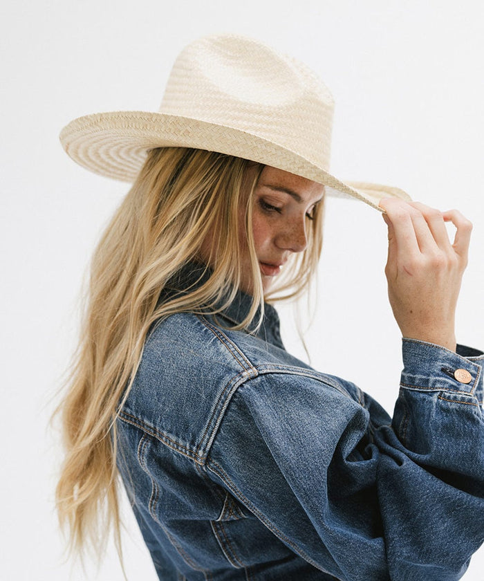 Woman wearing a natural colored straw cowboy hat and a denim shirt, posing sideways while holding her hat against a plain background  #color_natural