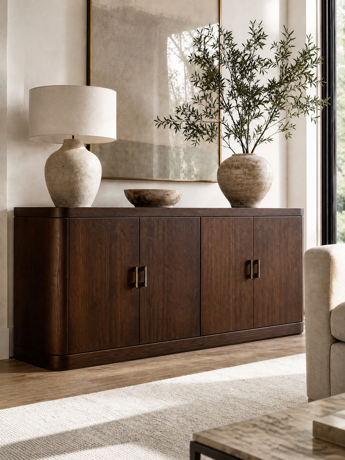 A dark brown wooden sideboard cabinet in a modern living room setting, styled with a textured ceramic table lamp, a small stone bowl, and a large vase with olive branches.