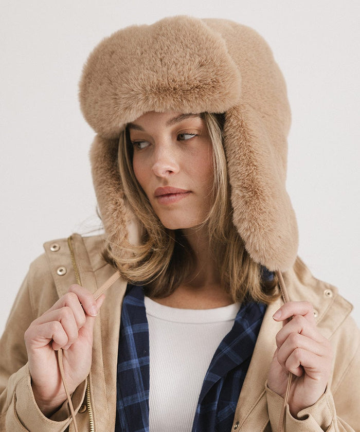 Woman wearing a tan fur hat with ear flaps and a beige jacket against a plain background #color_tan