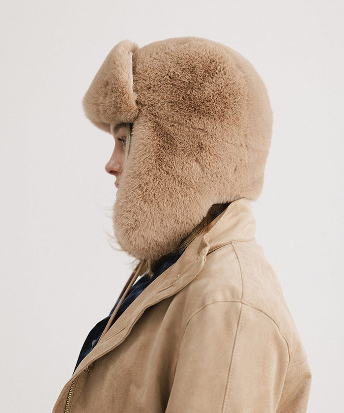 Woman wearing a tan fur hat with ear flaps and a beige jacket, facing sideways against a plain background #color_tan
