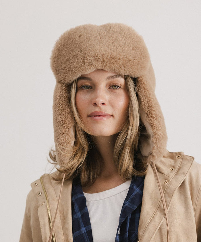 Woman wearing a tan fur hat with ear flaps and a white top with a beige jacket against a plain background #color_tan