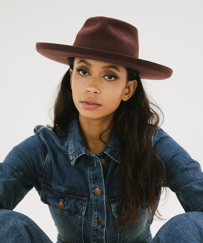 Woman wearing a dark cherry colored felt fedora hat and a denim shirt against a plain background #color_dark cherry
