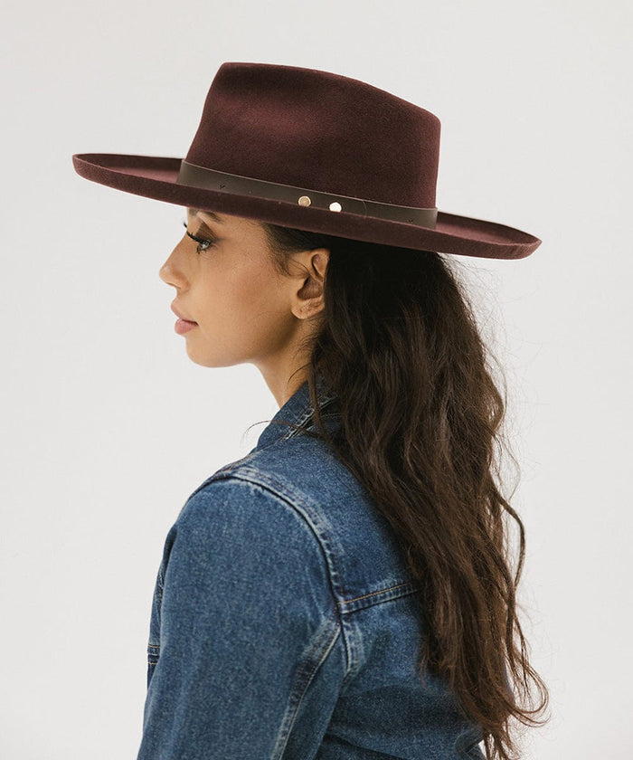 Woman wearing a dark cherry colored felt fedora hat with a hat band and a denim shirt, facing sideways against a plain background #color_dark cherry