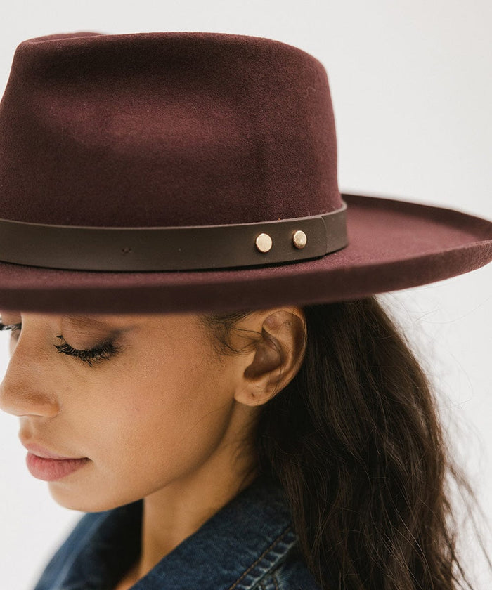 Woman wearing a dark cherry colored felt fedora hat with a hat band, facing sideways against a plain background #color_dark cherry
