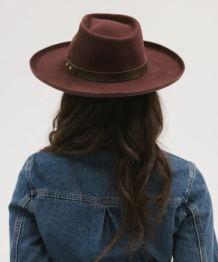 Person wearing a dark cherry colored felt fedora hat with a hat band and a denim shirt against a plain background #color_dark cherry