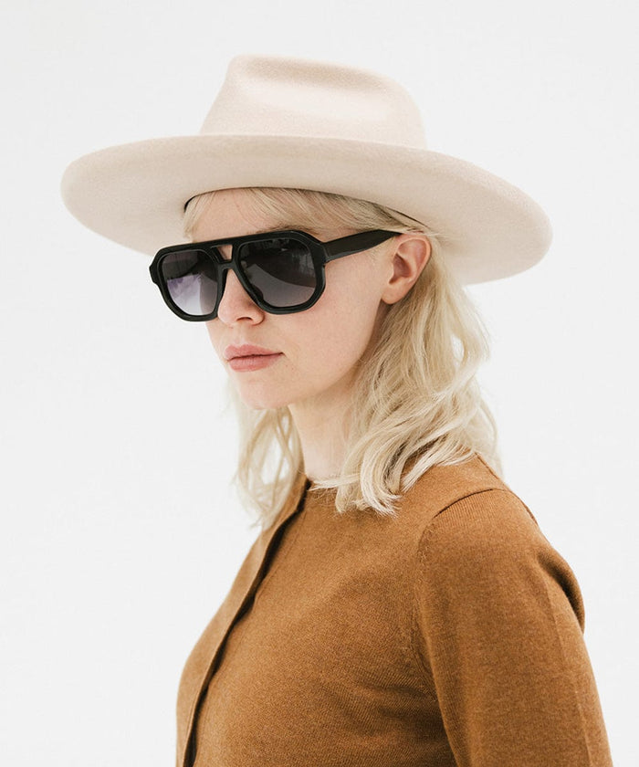 Woman wearing a cream felt fedora hat with a hat band, sunglasses, and a brown sweater against a plain background #color_cream