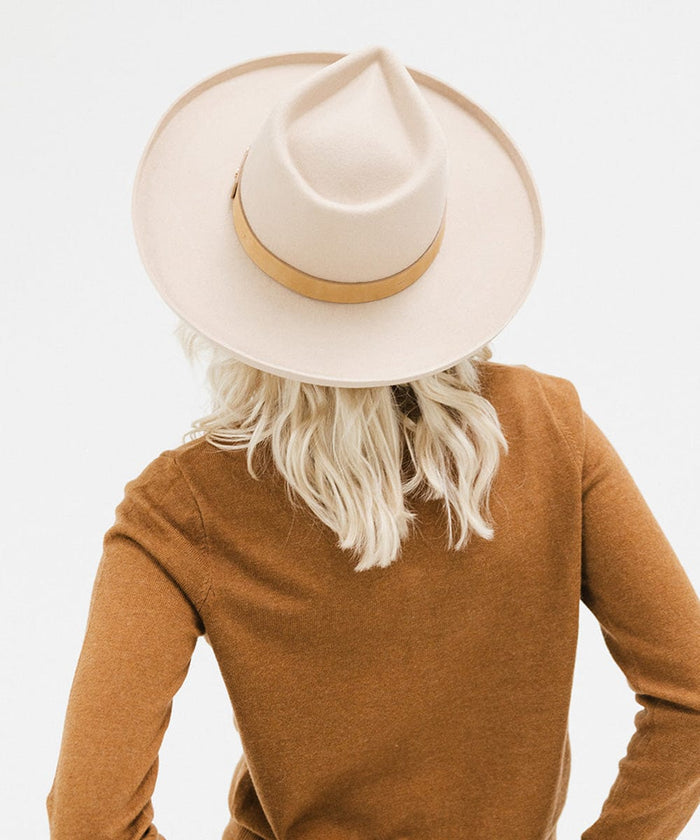 Woman wearing a cream felt fedora hat with a hat band and a brown sweater, facing behind against a plain background #color_cream