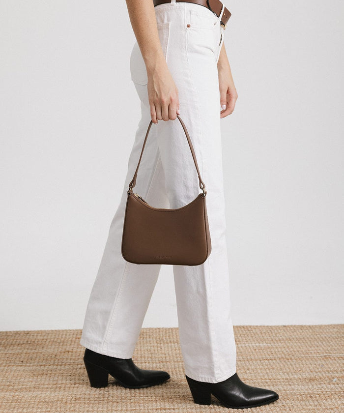 Person wearing a brown belt, white pants, and black shoes, carrying a chocolate colored genuine leather handbag against a plain background #color_chocolate