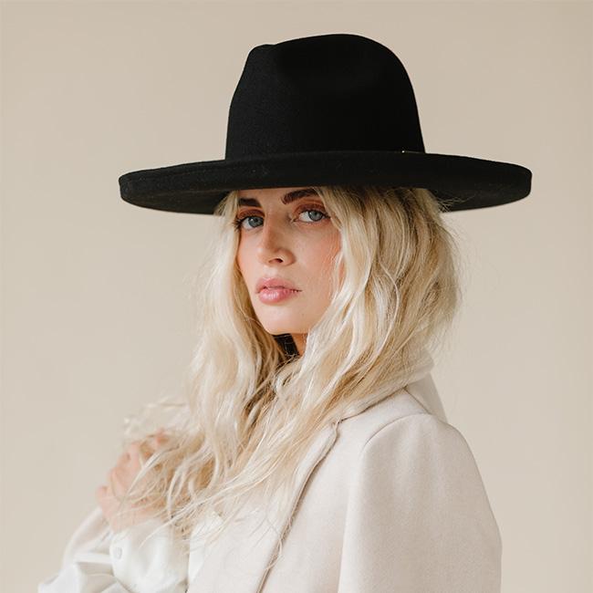 Woman wearing a black pencil brim felt hat and a white blazer against a plain background #color_black