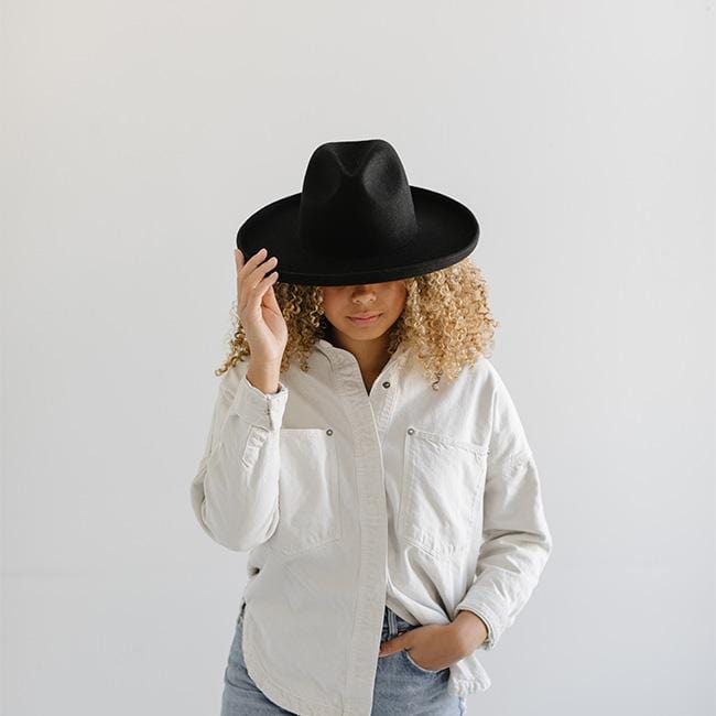 Woman wearing a black pencil brim felt hat, a white shirt and blue jeans against a plain background #color_black