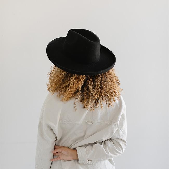 Woman wearing a black pencil brim felt hat and a white shirt, facing behind against a plain background #color_black