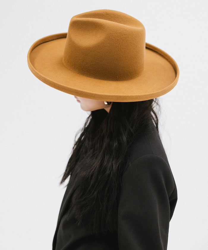Woman wearing a cinnamon colored  pencil brim felt fedora hat and a black blazer, facing sideways against a white background #color_cinnamon