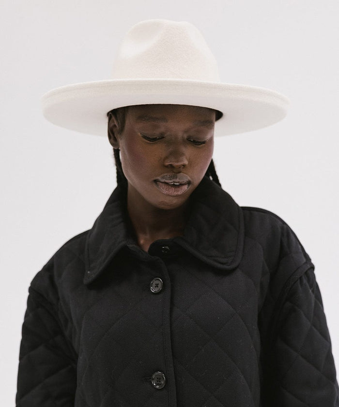 Woman wearing an off white pencil brim felt fedora hat and a black coat, looking down against a white background #color_off white