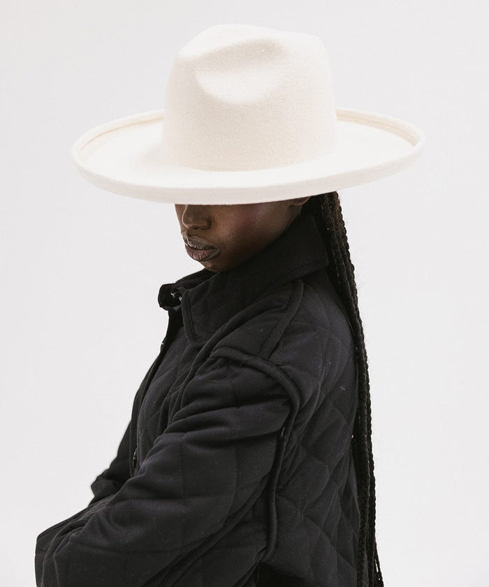 Woman wearing an off white pencil brim felt fedora hat and a black coat, facing sideways against a white background #color_off white