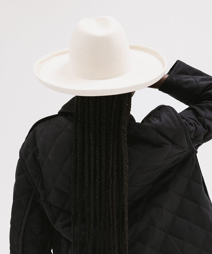 Woman wearing an off white pencil brim felt fedora hat and a black coat, facing behind against a white background #color_off white
