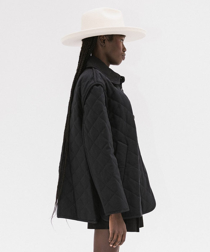 Woman wearing an off white pencil brim felt fedora hat and a black coat, facing sideways against a white background #color_off white