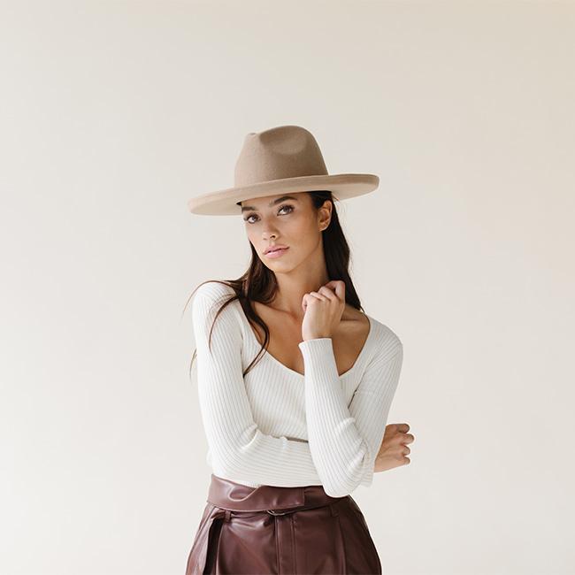 Woman wearing a tan pencil brim fedora hat, a white top and brown skirt against a plain background #color_tan