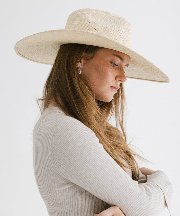 Woman wearing a natural colored wide brim straw hat and a white top, facing sideways against a plain background  #color_natural