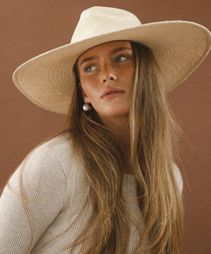 Woman wearing a natural colored wide brim straw hat and a white top against a brown background  #color_natural