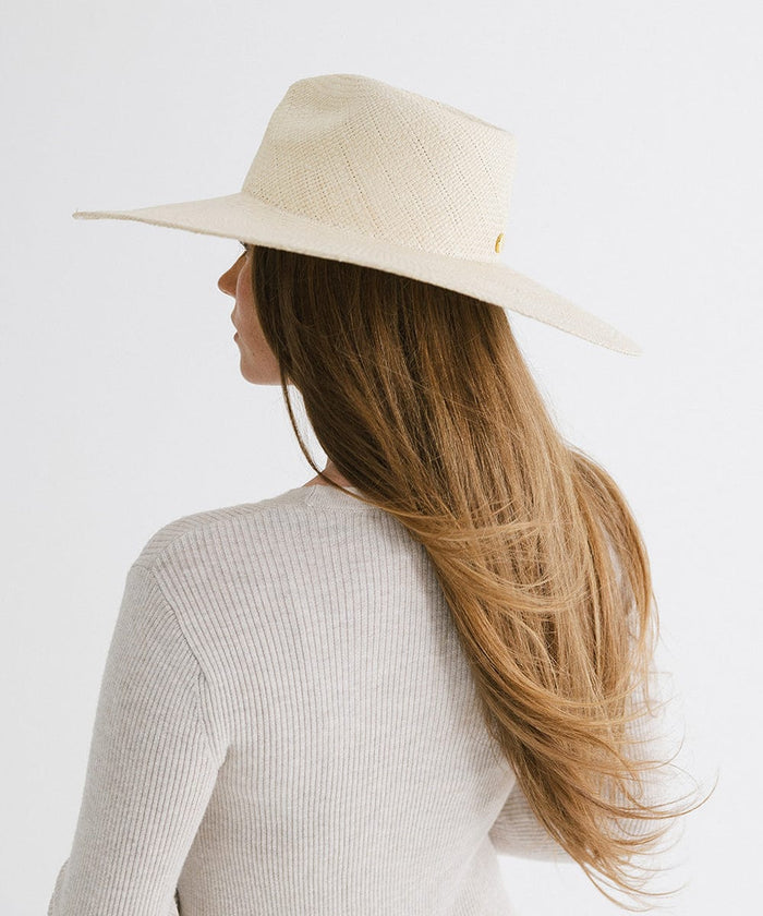 Woman with long hair wearing a natural colored wide brim straw hat and a white top, facing behind against a plain background  #color_natural