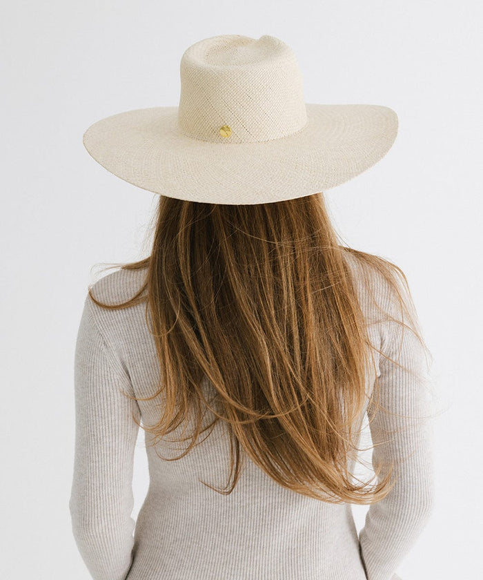 Person with long hair wearing a natural colored wide brim straw hat with a gold pin and a white top, facing behind against a plain background  #color_natural
