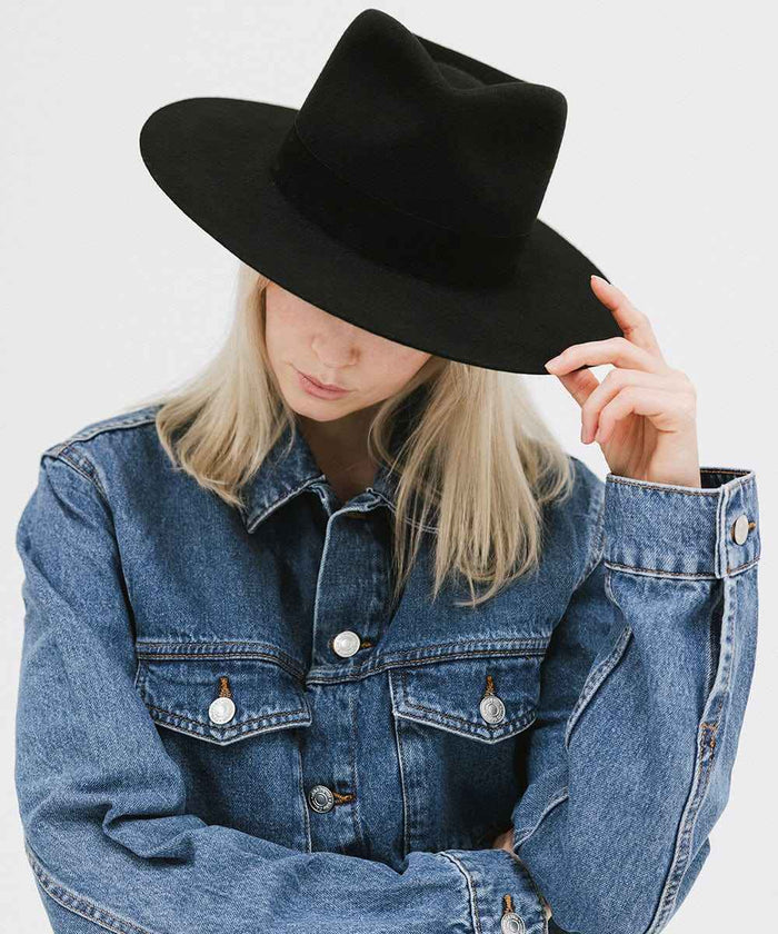 Person wearing a black fedora hat with a band and blue denim jacket on a white background #color_black