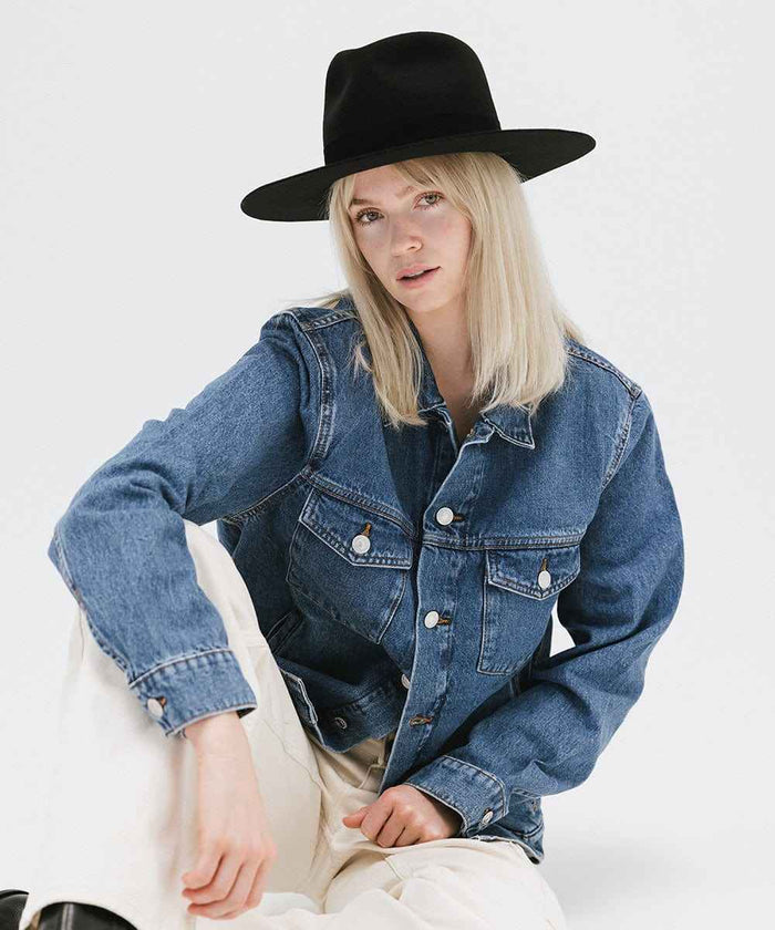 Person wearing a black fedora hat with a band, a denim jacket and white pants on a white background #color_black