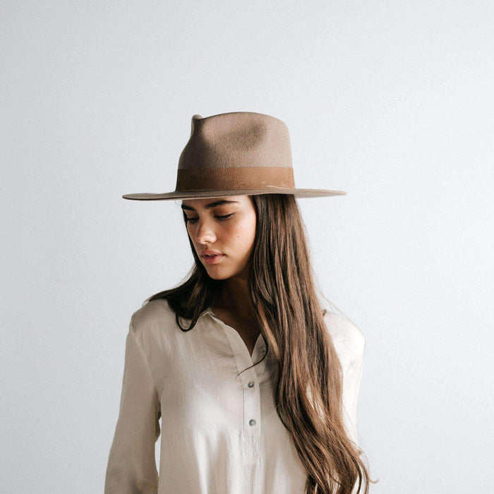Woman with long hair wearing a brown fedora hat with a band and a white shirt against a plain background #color_brown