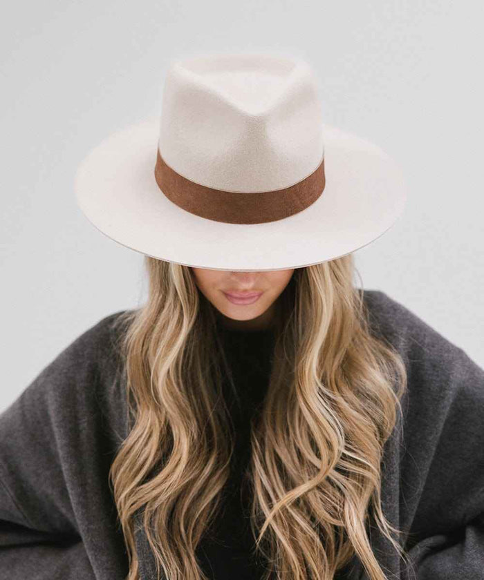 Woman wearing an ivory colored fedora hat with a brown band and a dark grey coat on a light grey background  #color_ivory