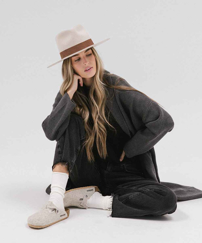 Woman wearing a dark grey coat, an ivory colored fedora hat, black jeans and white shoes, sitting against a white background #color_ivory