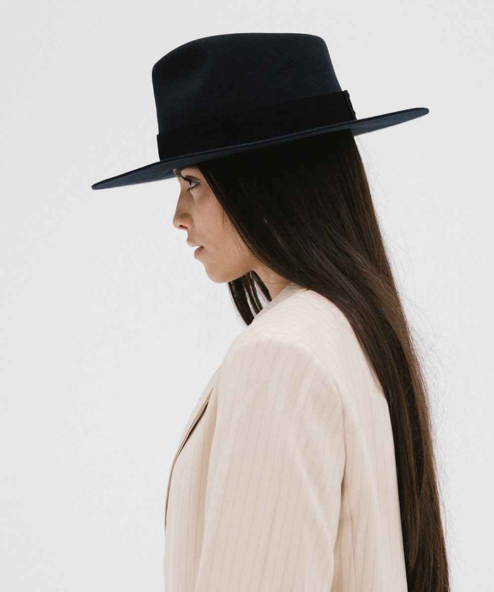 Woman with long hair wearing a navy fedora hat with a band and beige jacket, facing sideways against a white background #color_navy