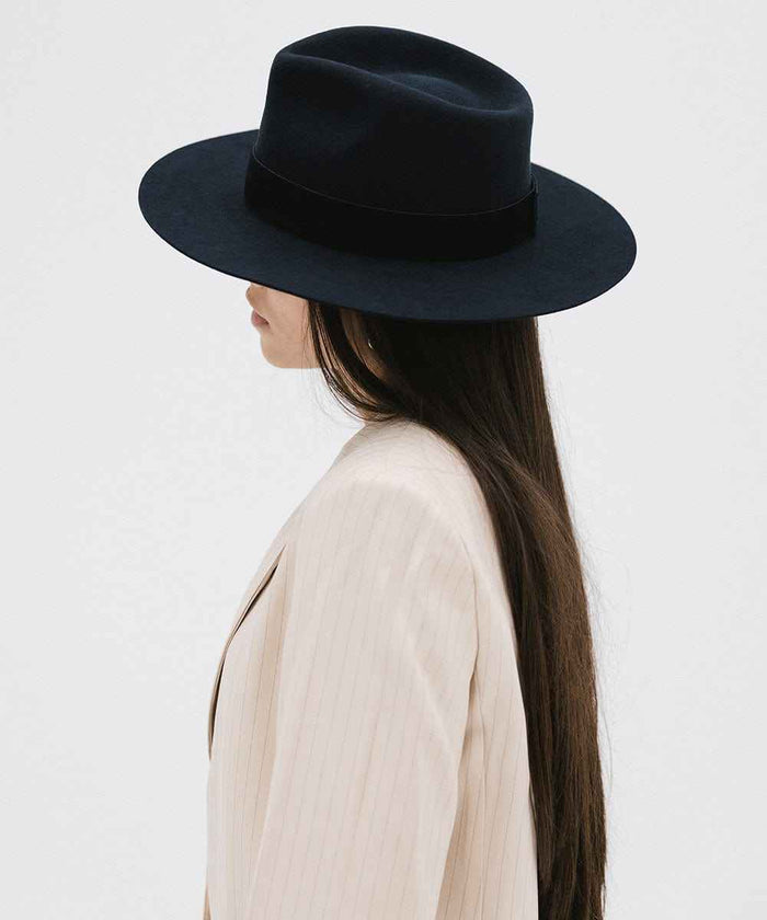 Woman wearing a navy fedora hat with a band and a beige blazer, looking sideways against a white background #color_navy
