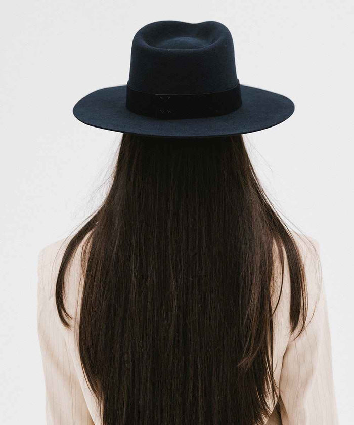 Woman with long hair wearing a navy fedora hat with a band facing behind against a plain background #color_navy
