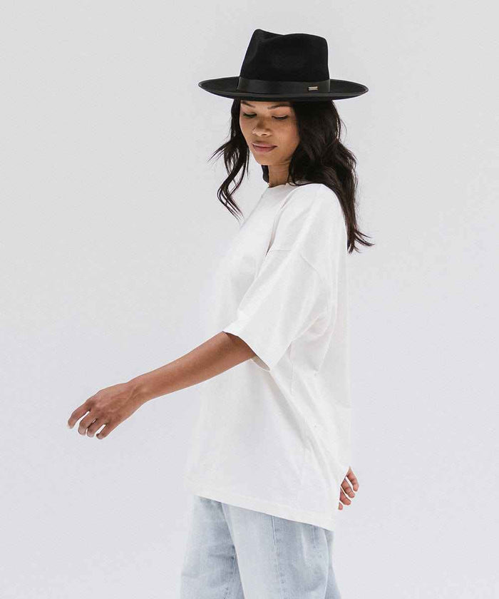 Woman wearing a black fedora hat with a band, a white t-shirt and light blue jeans against a plain background #color_black