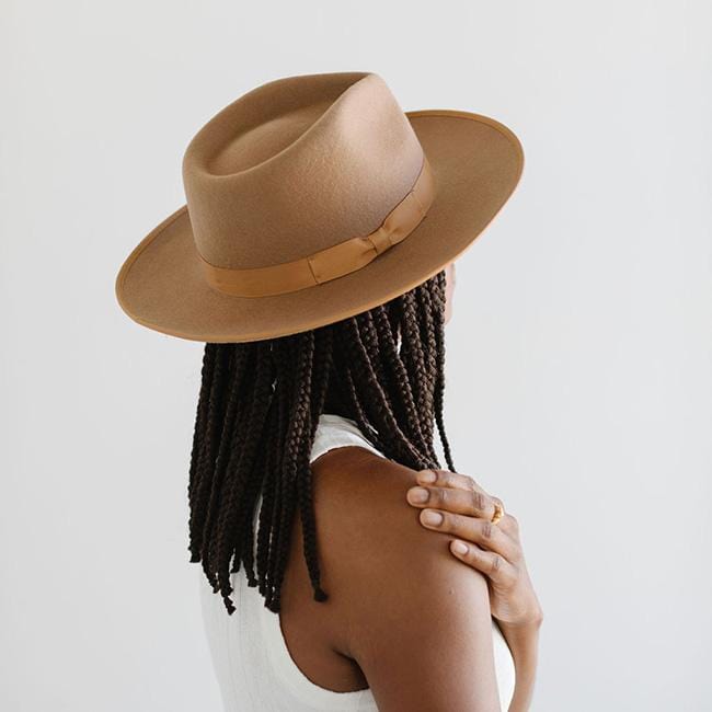 Woman wearing a brown fedora hat with a hat band and a white sleeveless top, facing behind against a plain background #color_brown