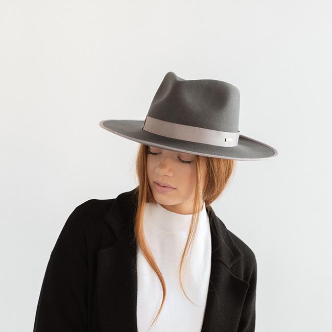 Woman wearing a dark grey fedora hat with a band and a white t-shirt with a black jacket against a plain background #color_dark grey