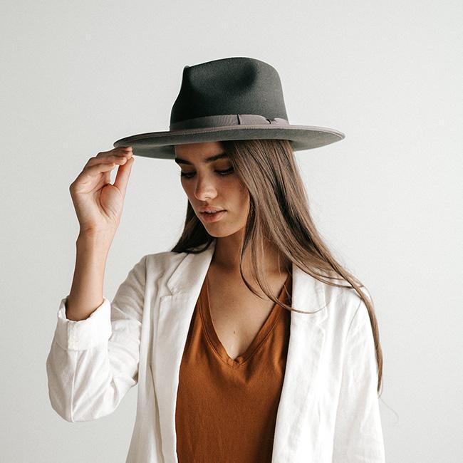 Woman wearing a dark grey fedora hat with a band and a brown t-shirt with a white jacket against a plain background #color_dark grey
