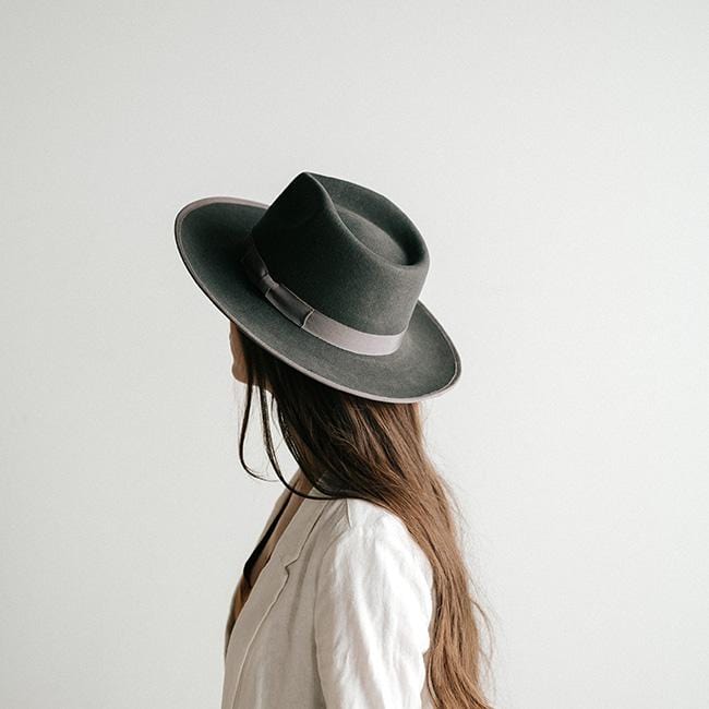 Woman wearing a dark grey fedora hat with a band and a white jacket, looking sideways against a plain background #color_dark grey