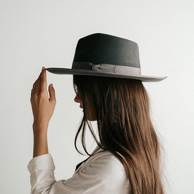 Woman wearing a dark grey fedora hat with a band and a white jacket, facing sideways against a plain background #color_dark grey