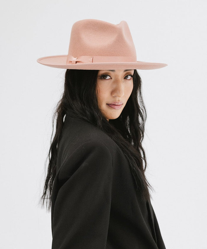 Woman wearing a dusty pink colored fedora hat with a band and a black blazer, posing against a plain background #color_dusty pink
