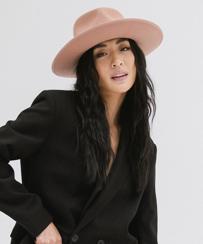 Woman wearing a dusty pink colored fedora hat with a band and a black blazer against a plain background #color_dusty pink