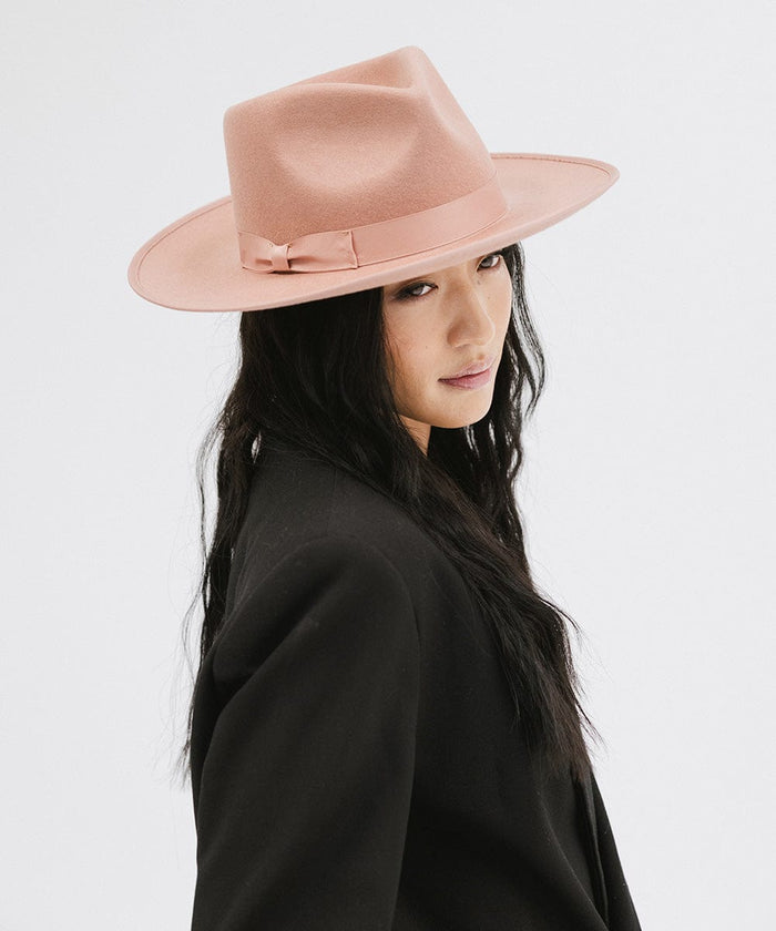 Woman wearing a dusty pink colored fedora hat with a band and a black blazer, looking sideways against a plain background #color_dusty pink