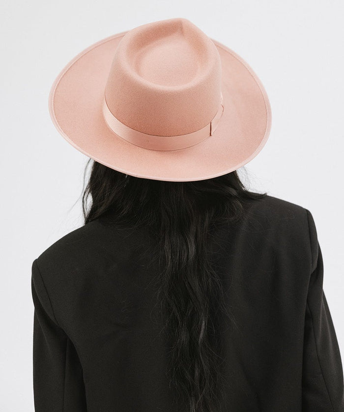 Woman wearing a dusty pink colored fedora hat with a band and a black blazer, facing behind against a plain background #color_dusty pink