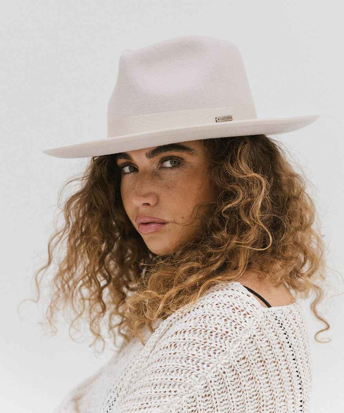 Woman wearing an ivory colored fedora hat with a band and a white knit sweater, posing against a plain background #color_ivory