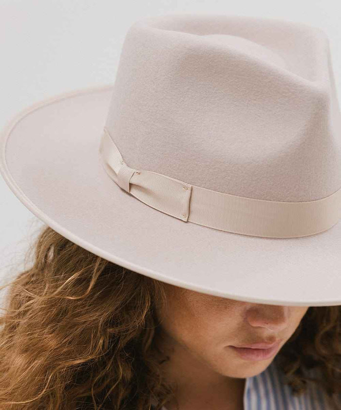 Woman wearing an ivory colored fedora hat with a band against a plain background #color_ivory