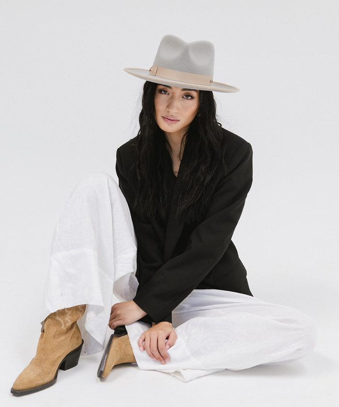 Woman wearing a light grey and tan colored fedora hat with a tan band, a black blazer, white pants and brown boots, sitting against a plain background #color_light grey-tan
