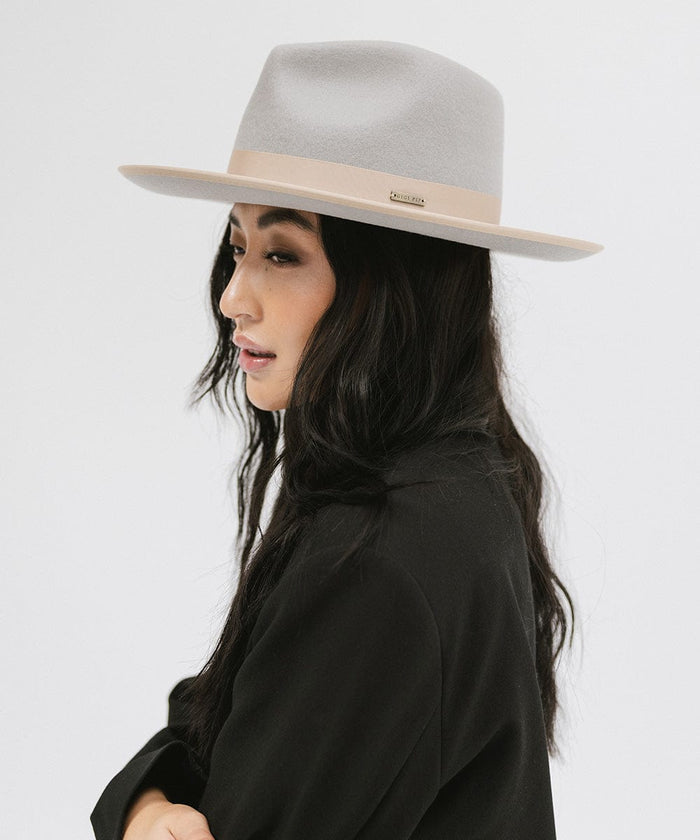 Woman wearing a light grey and tan colored fedora hat with a tan band and a black blazer, looking sideways against a plain background #color_light grey-tan