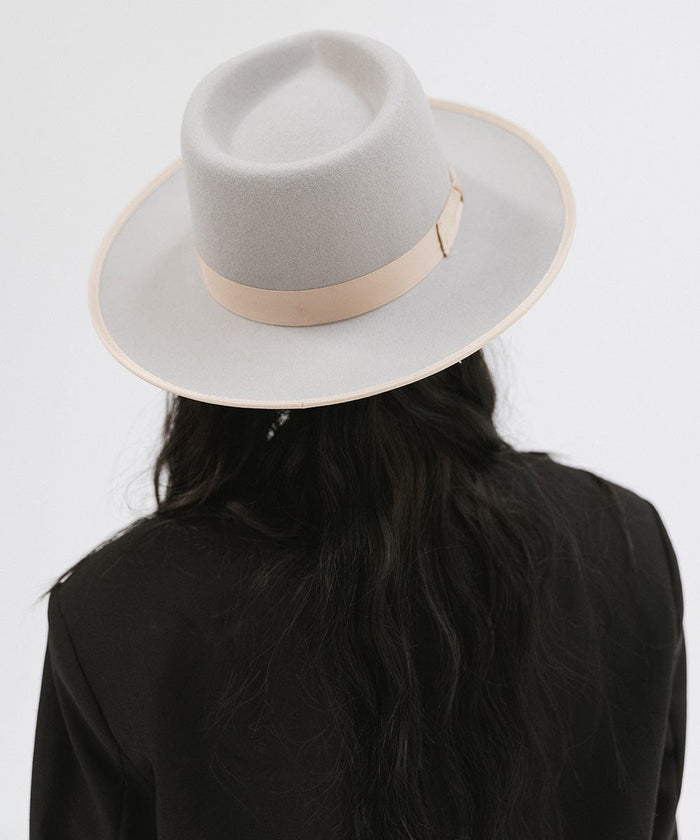 Woman wearing a light grey and tan colored fedora hat with a tan band and a black blazer, facing behind against a plain background #color_light grey-tan